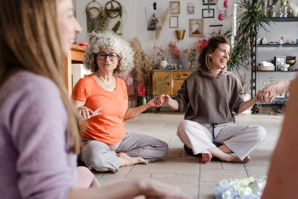 Mehrere Teilnehmerinnen von Sonja Blessings Massageausbildung bilden hier einen Sitzkreis und reichen sich lachend die Hände.